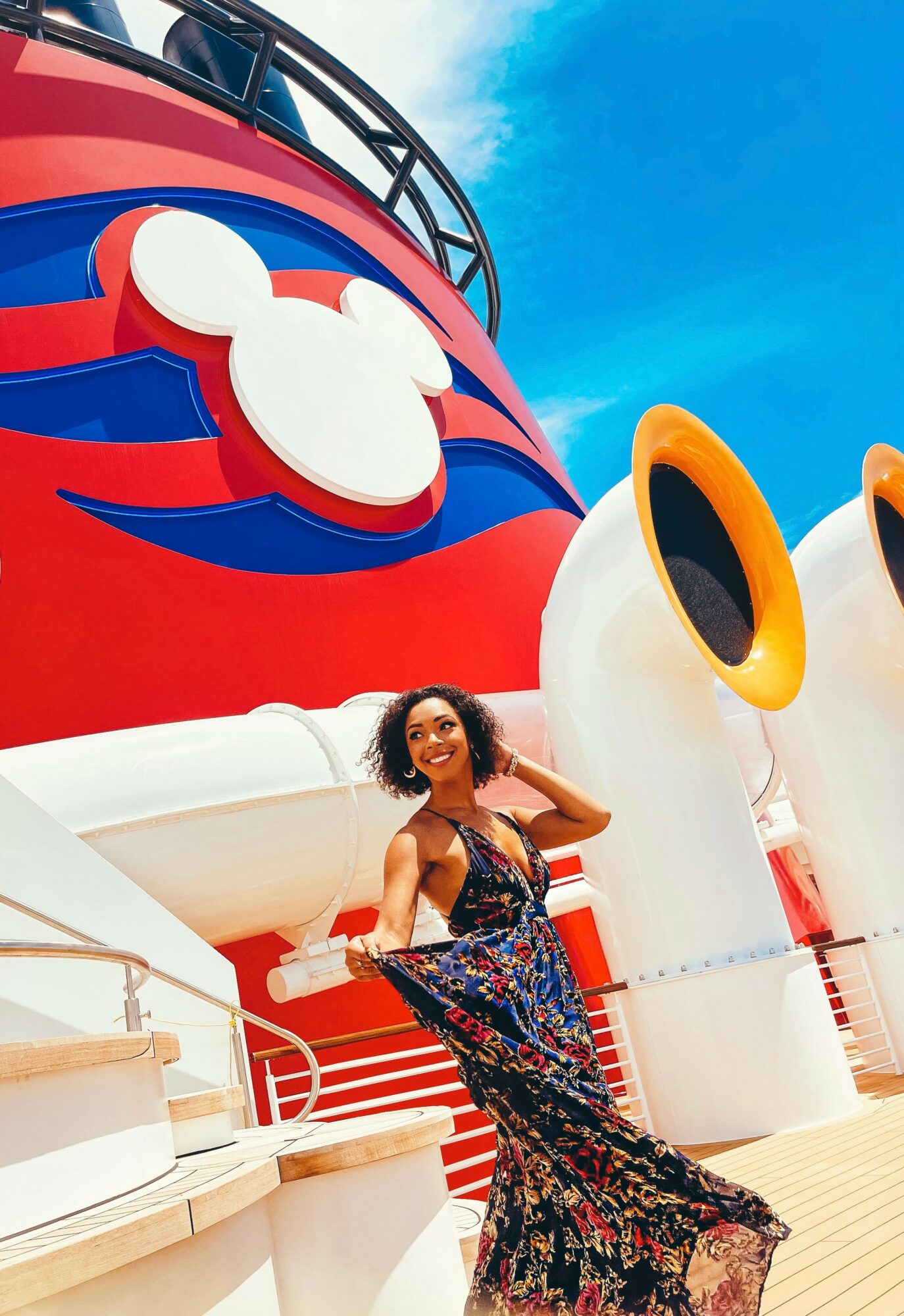Woman in colorful dress standing in front of a large Disney-themed structure with a Mickey Mouse logo, smiling, outdoors on a sunny day.
