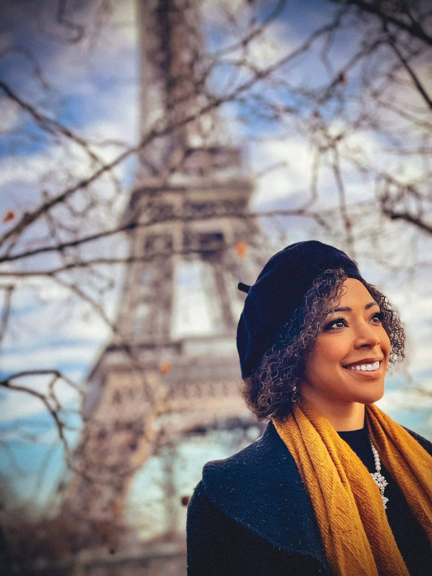 Young woman smiling outdoors with the Eiffel Tower in the background, wearing a black beret and yellow scarf.