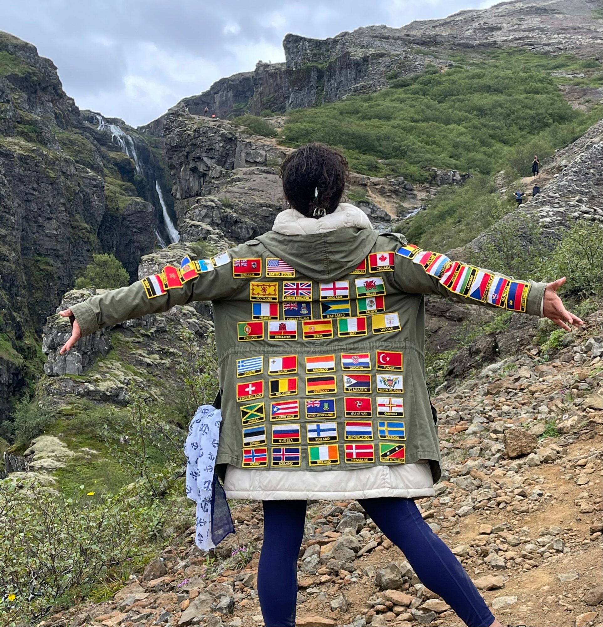 Person standing outdoors with arms outstretched, back facing camera, wearing a jacket decorated with numerous international flags, mountainous landscape in background.