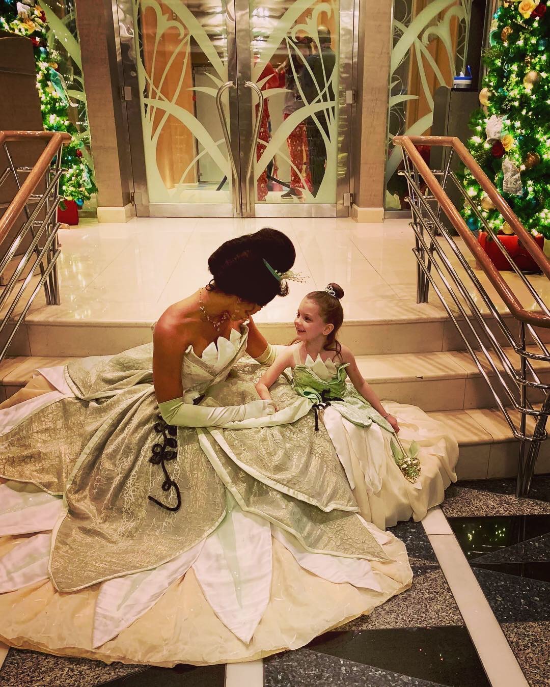 Two women in elegant dresses sitting on a staircase, with a decorated background and glass doors behind them.