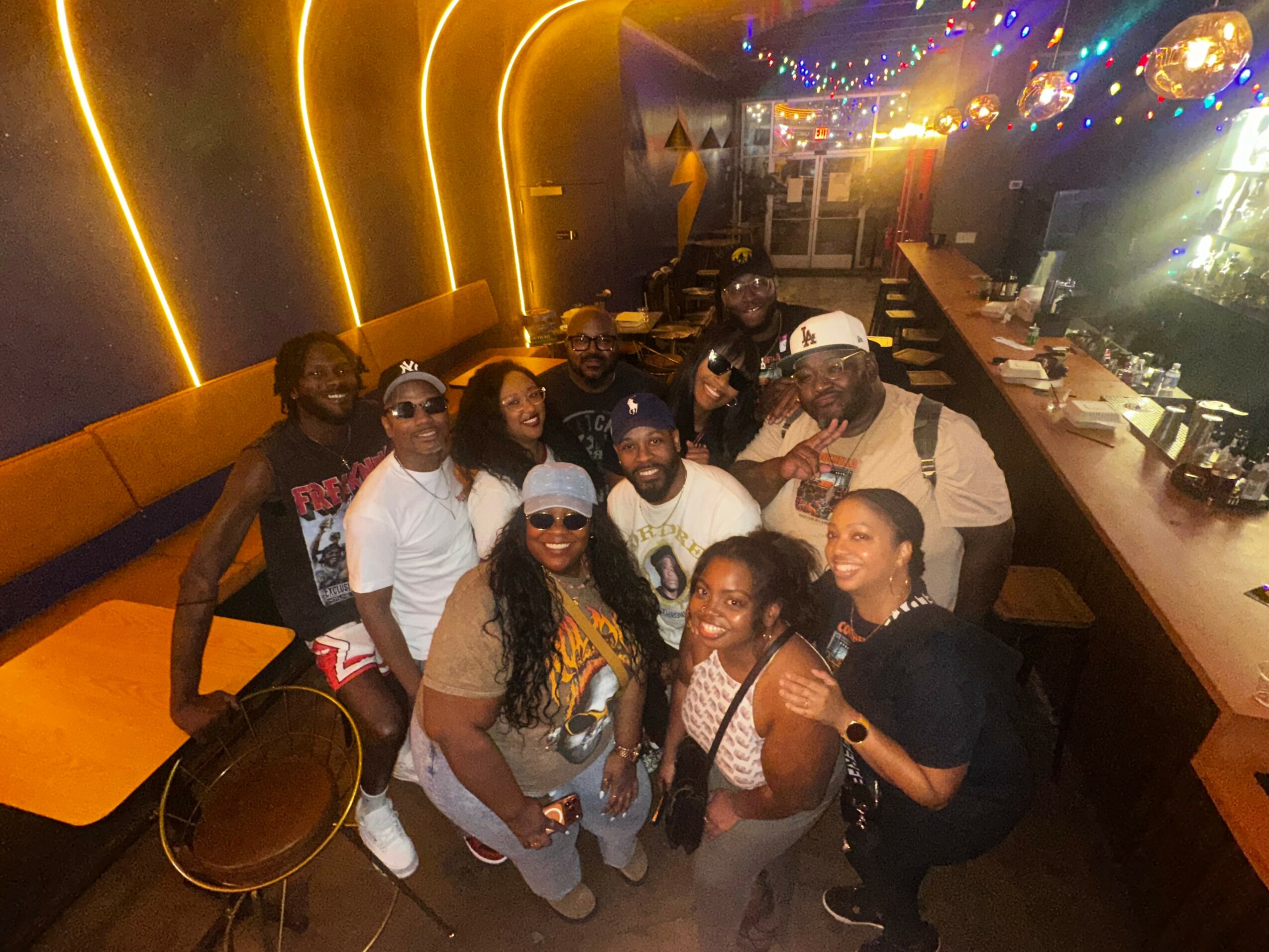 Group of people smiling and posing in a dimly lit restaurant or bar with warm lighting and a bar counter in the background.
