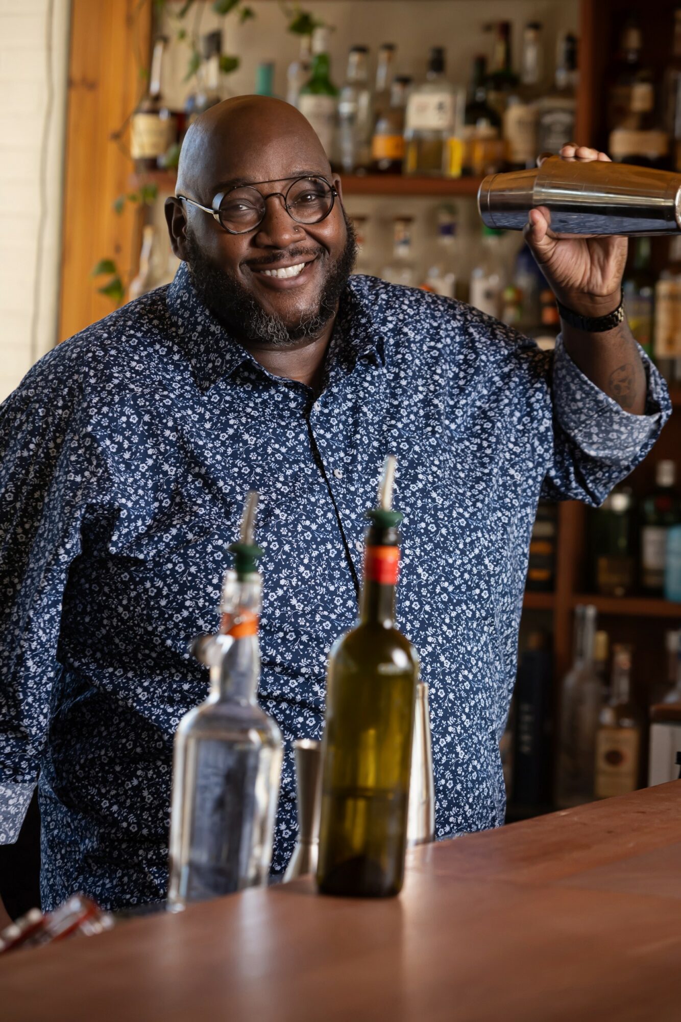 Smiling man with glasses behind bar, holding a book, with bottles on the counter and shelves in background.