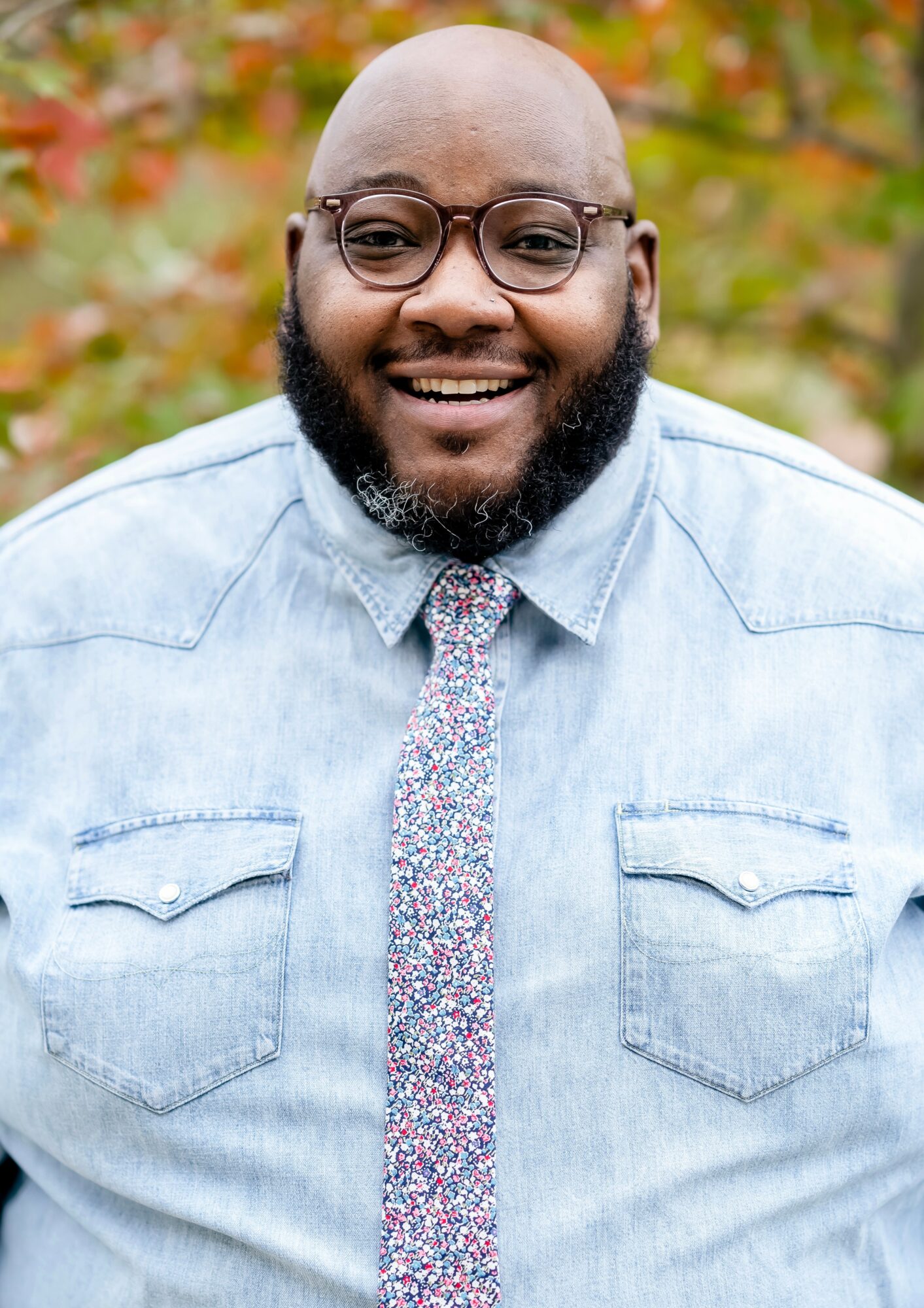 Smiling man with glasses and a beard, wearing a light denim shirt and a floral tie, outdoors with autumn foliage