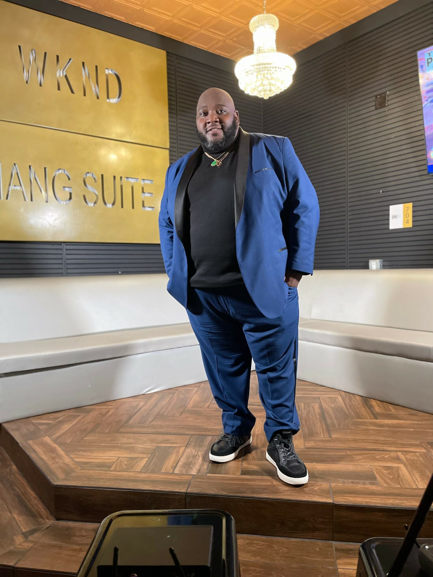 Man in blue suit standing in a room with a chandelier and gold sign, smiling, with a white bench behind him.
