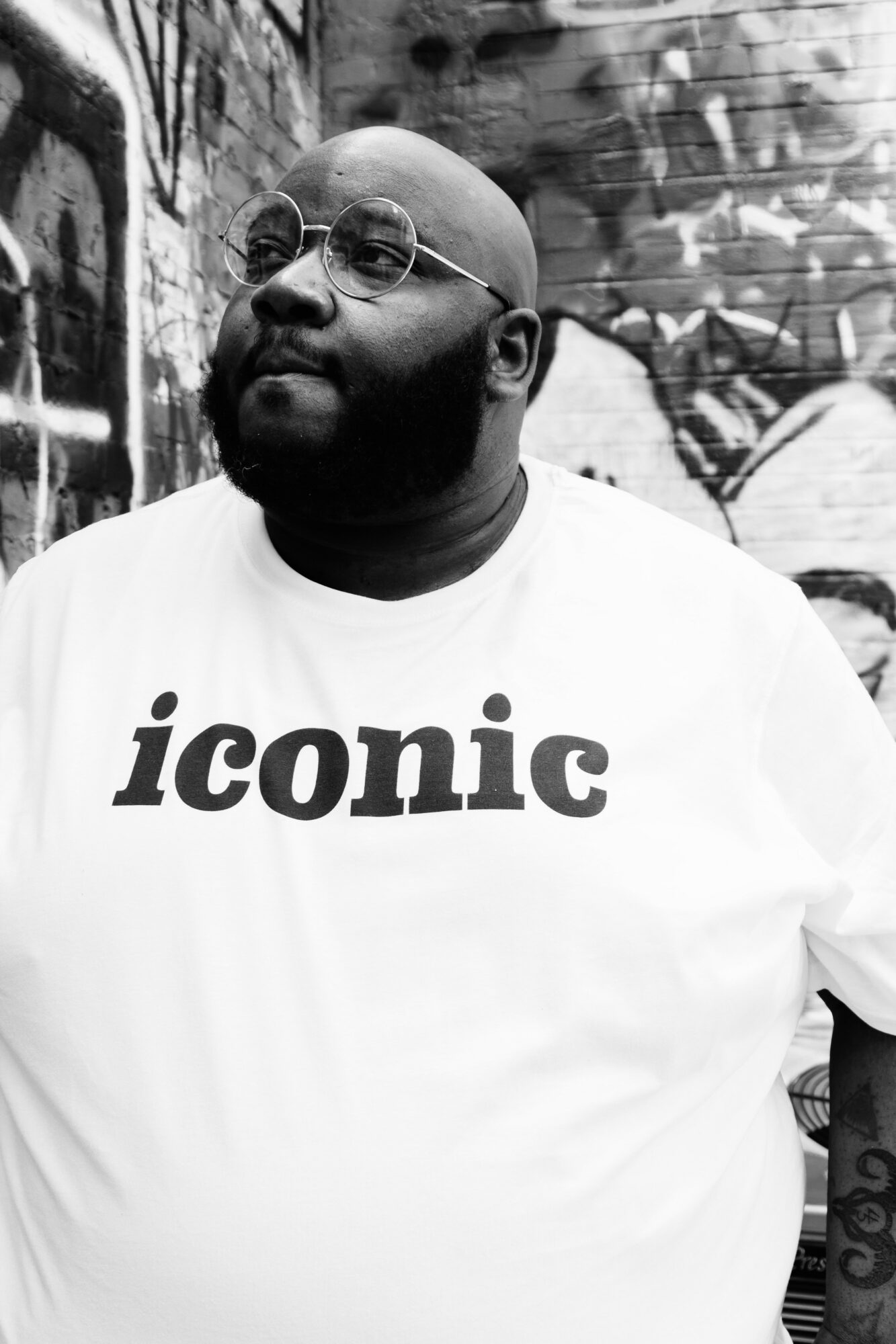 Man with glasses and beard wearing a white shirt with 'iconic' text, standing in front of graffiti wall.