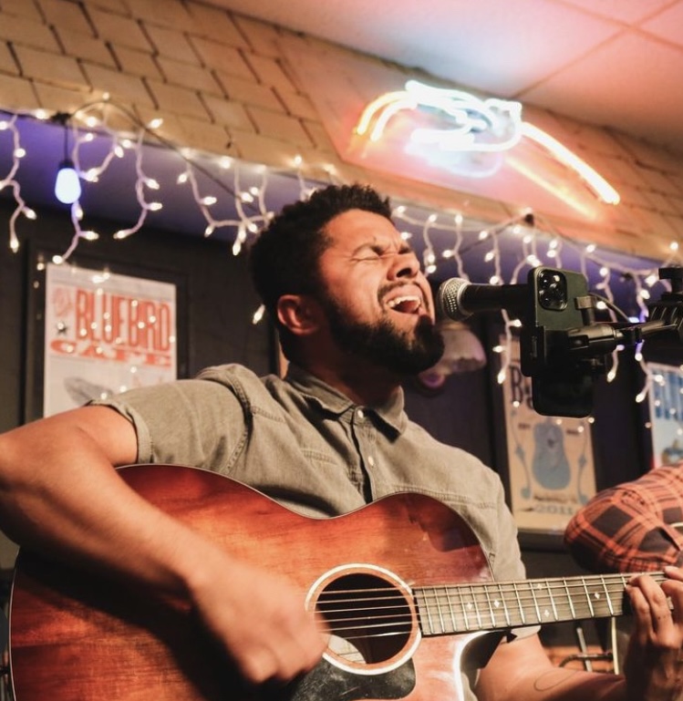 Man passionately singing and playing guitar in a decorated indoor setting with string lights and posters.