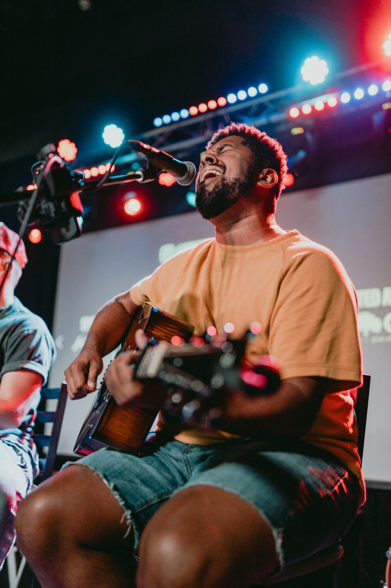 Man with a beard singing and playing guitar on stage with colorful lights behind him.