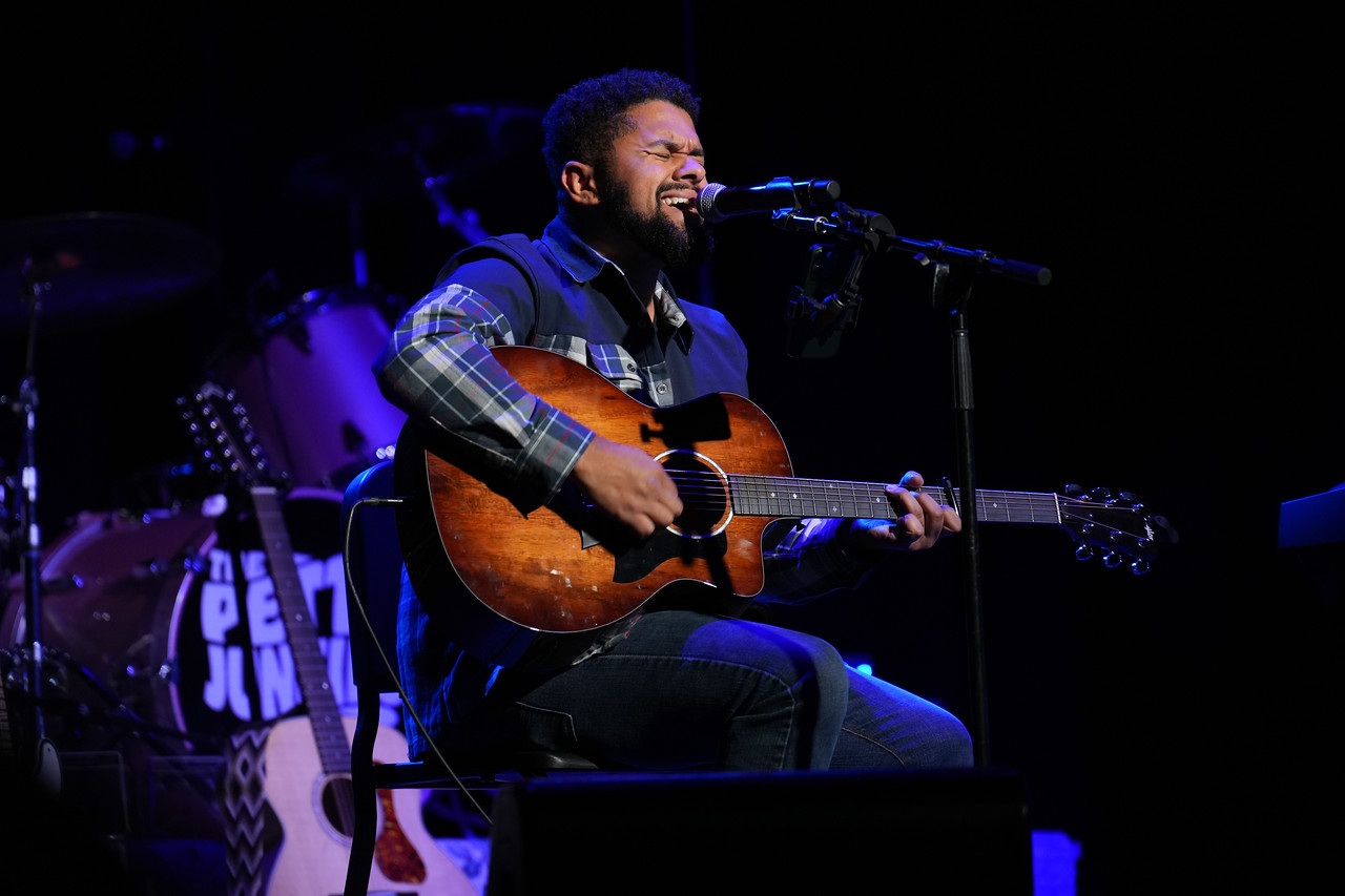Man playing guitar and singing on stage with microphone, dark background, purple lighting, smiling, performing live.