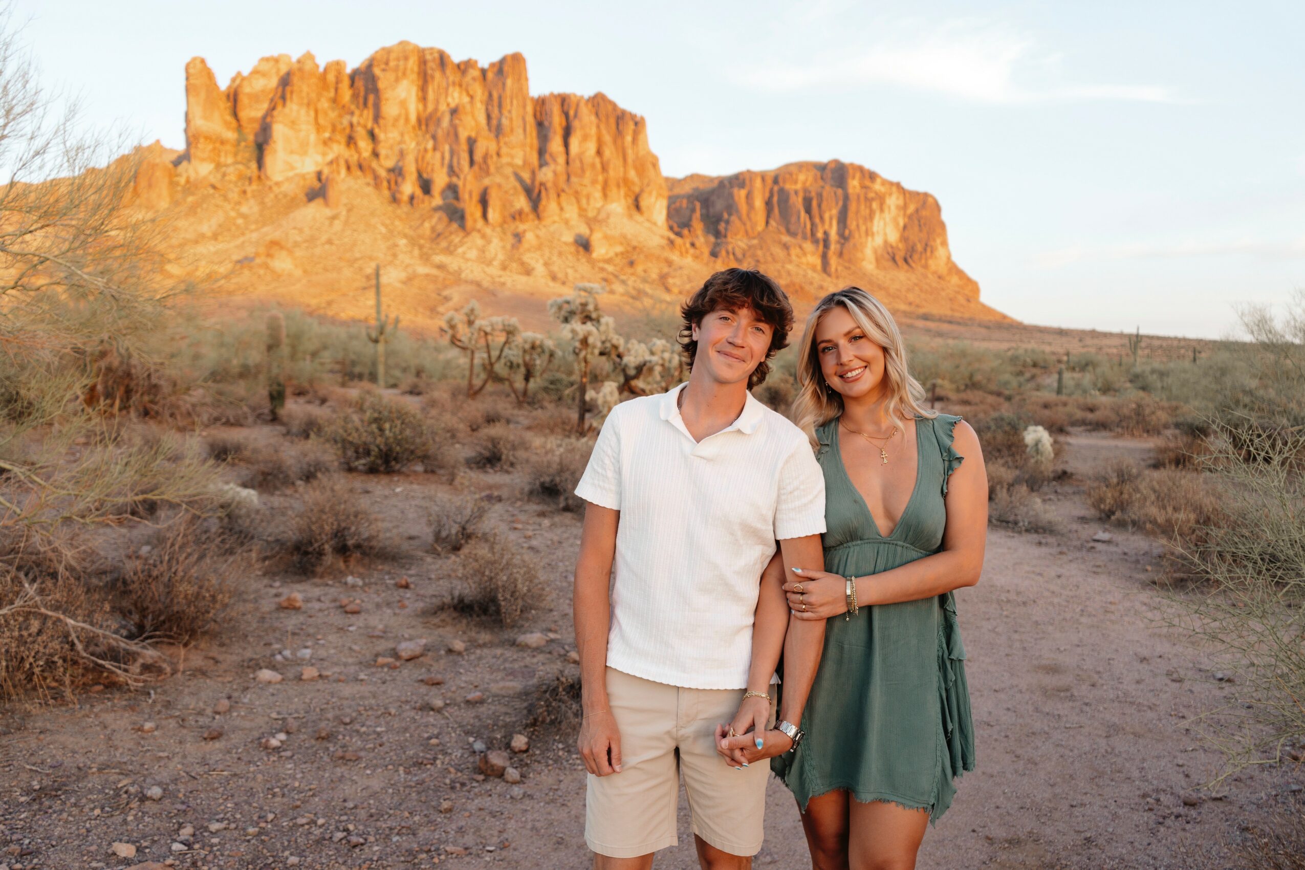 A young man and woman stand together in a desert landscape with a rocky formation in the background, smiling.