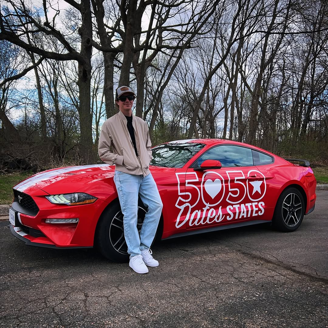Person standing next to a red sports car with white text and symbols, trees in the background.