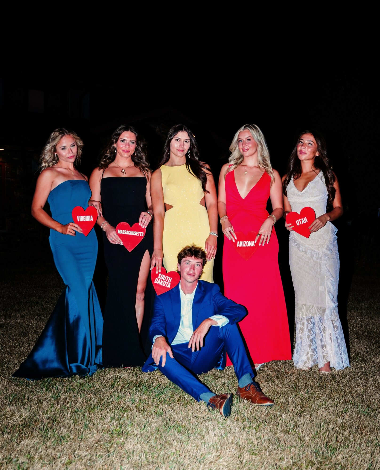 Group of five women and one man in formal dresses and suit standing outdoors at night, holding red heart-shaped signs.