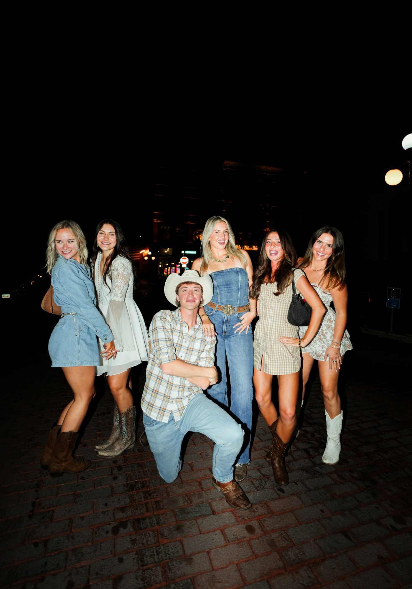 Group of six women and one man standing outdoors at night, smiling, with dark background and streetlights, brick ground.
