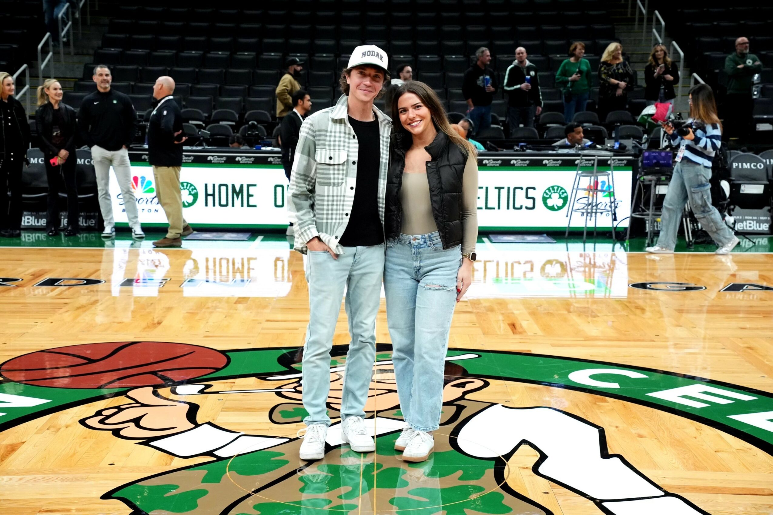 Two people stand on a basketball court with a large logo, smiling at the camera, with others in the background.