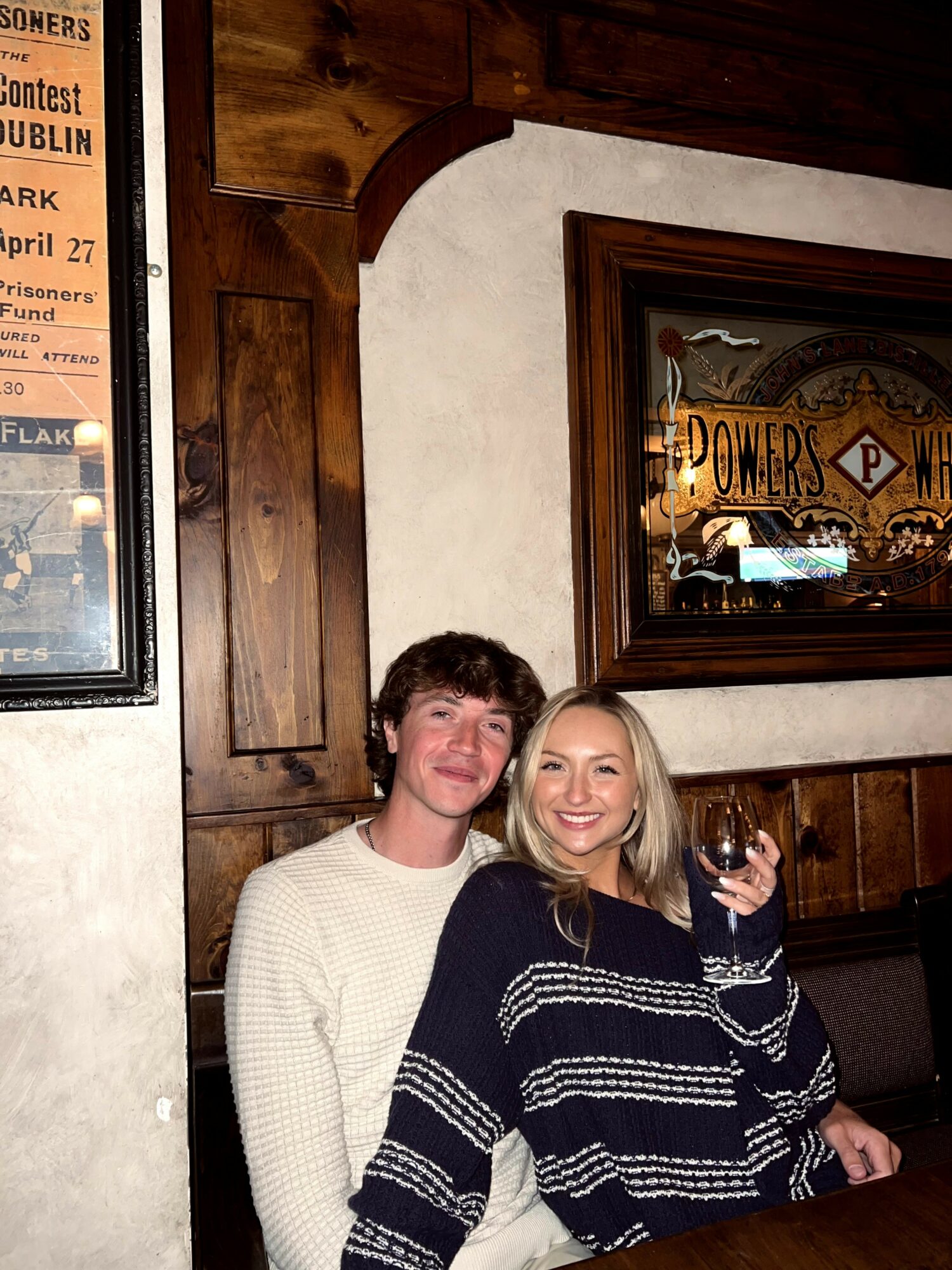 Two smiling people sitting close together in a wooden-paneled room, one holding a drink, with framed artwork on the wall behind them.
