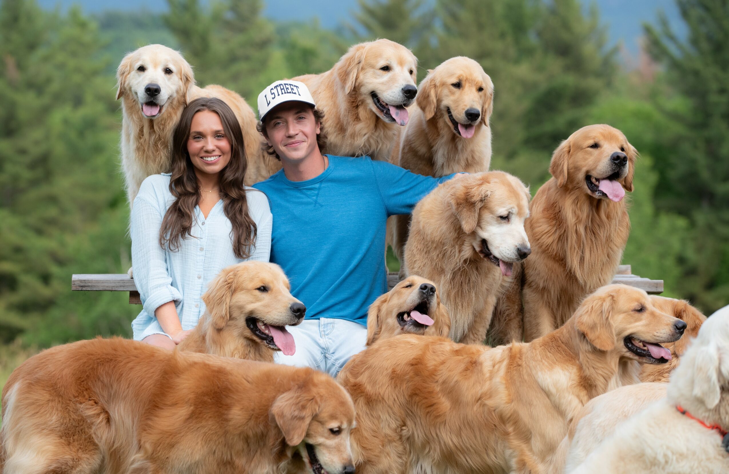 Two people and multiple golden retrievers outdoors with trees in the background.