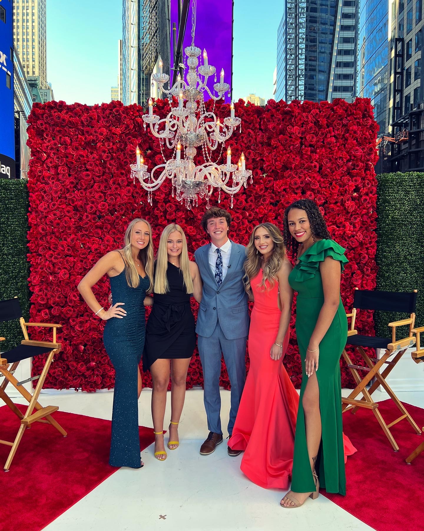 Group of five people in formal attire standing on a red carpet in front of a large red flower wall with a chandelier above, surrounded by chairs.