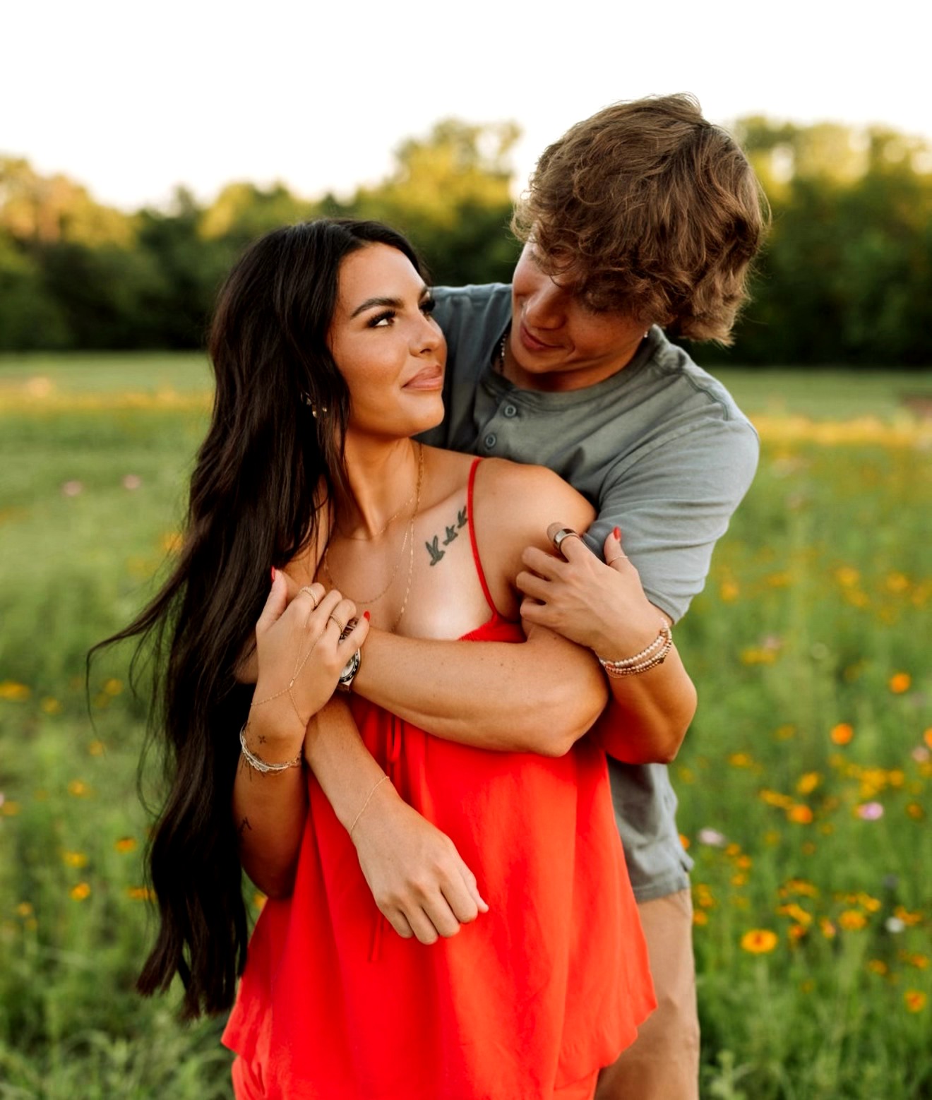 A man and woman embrace outdoors in a grassy field with yellow flowers, trees, and a blue sky in the background.