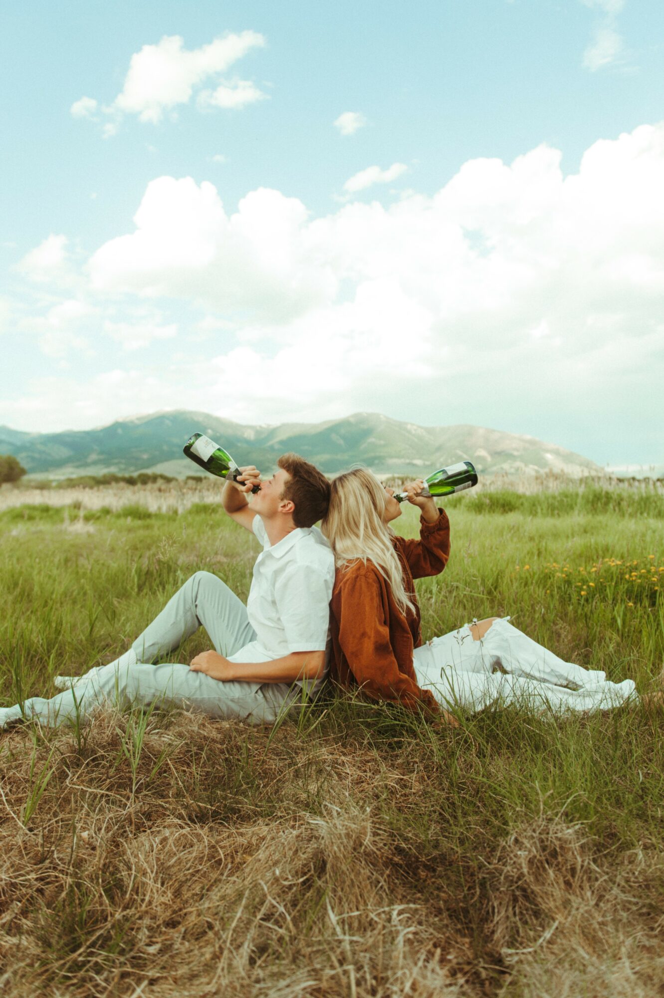 Two people sit on grass in a field, drinking from bottles, with a landscape and sky in the background.