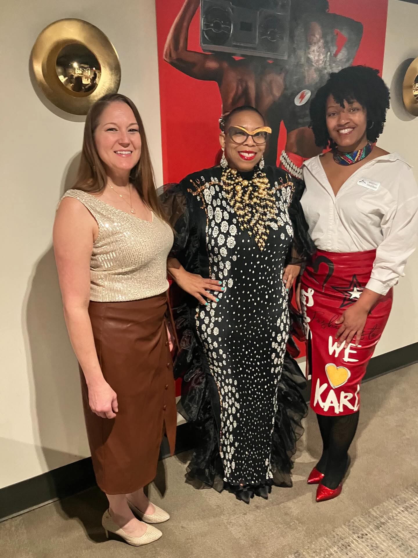 Three women standing together indoors, smiling, with a red and black wall art behind them.