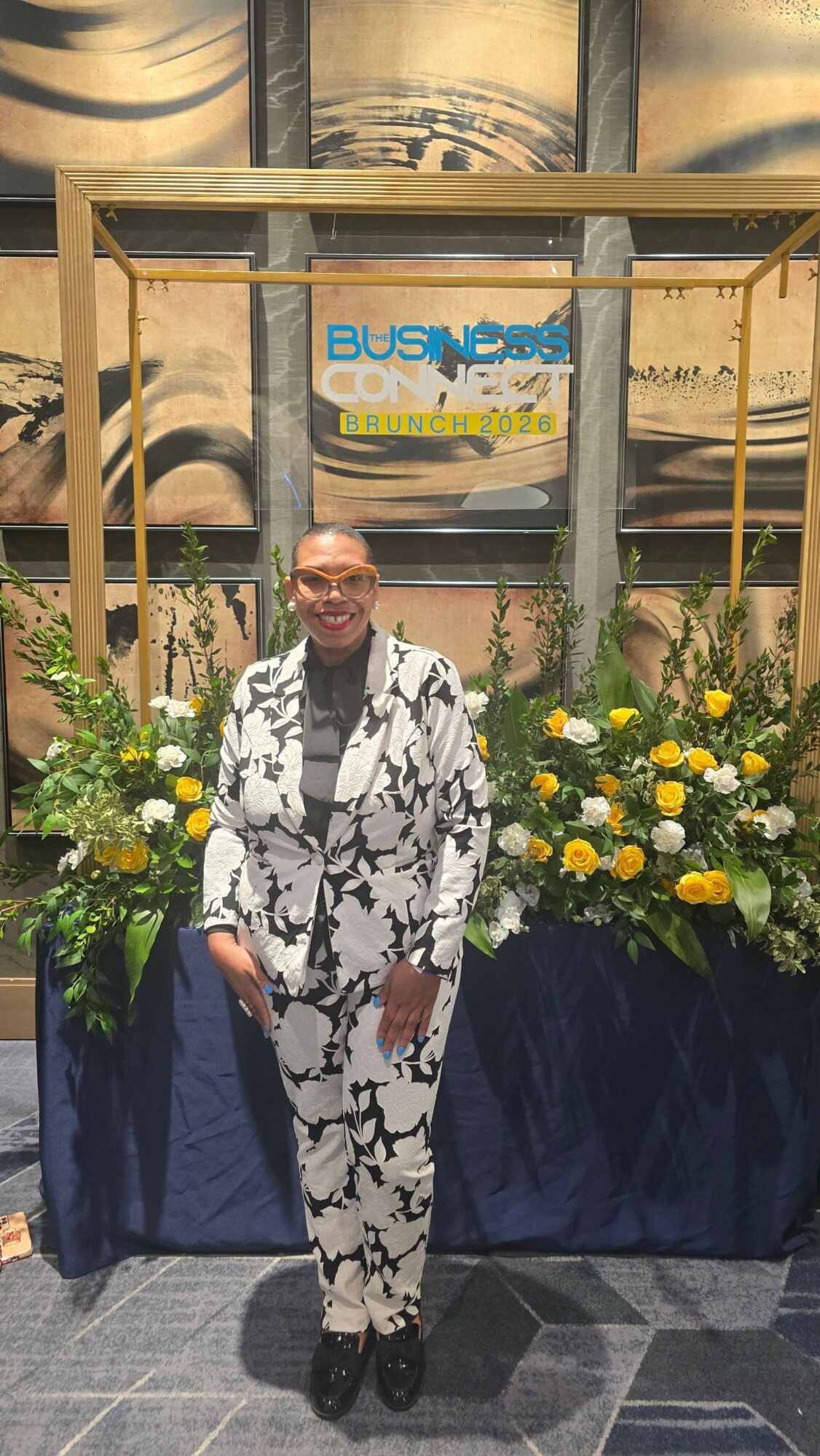 Person standing outdoors near a bus stop with yellow flowers and greenery, wearing a patterned outfit, smiling.