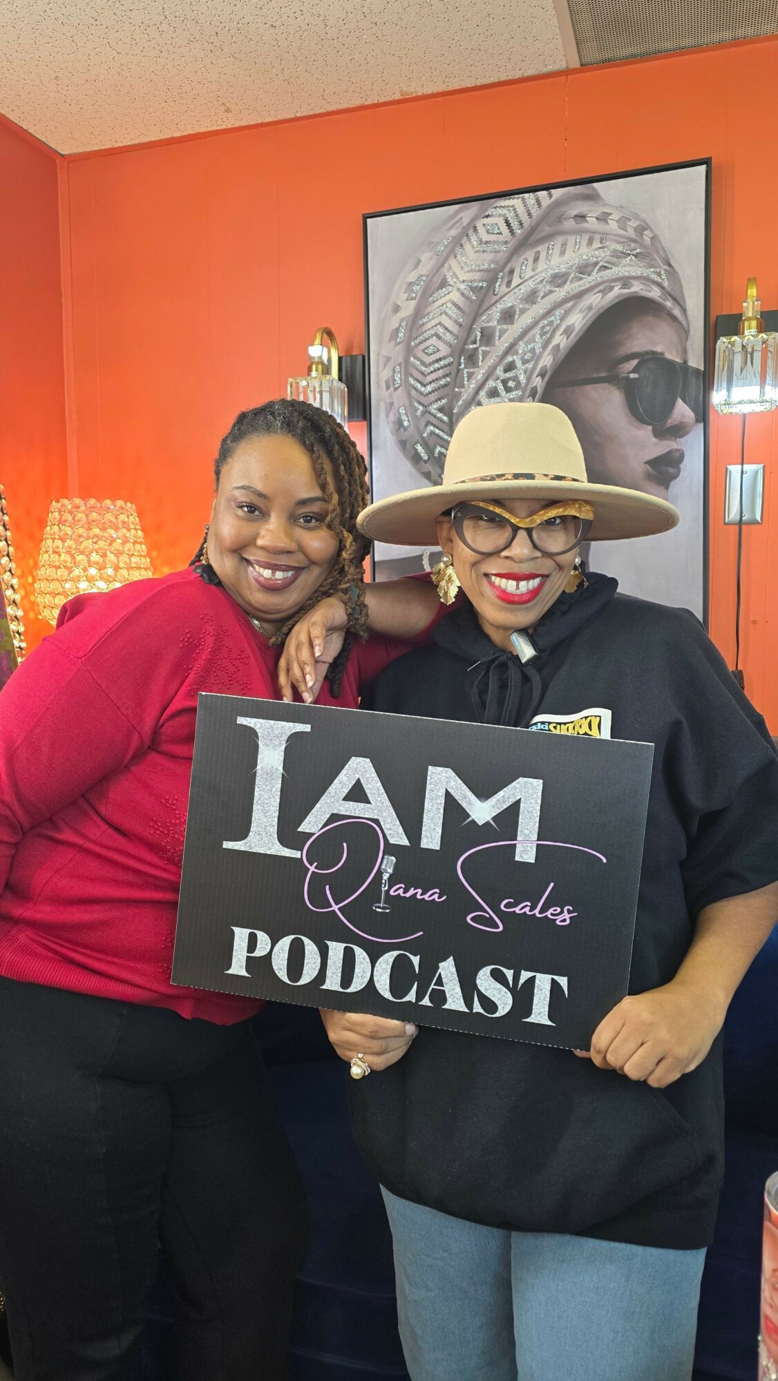 Two women sitting together, one holding a sign that reads 'I AM an LGBTQ PODCAST,' in front of an orange wall with a black-and-white portrait.