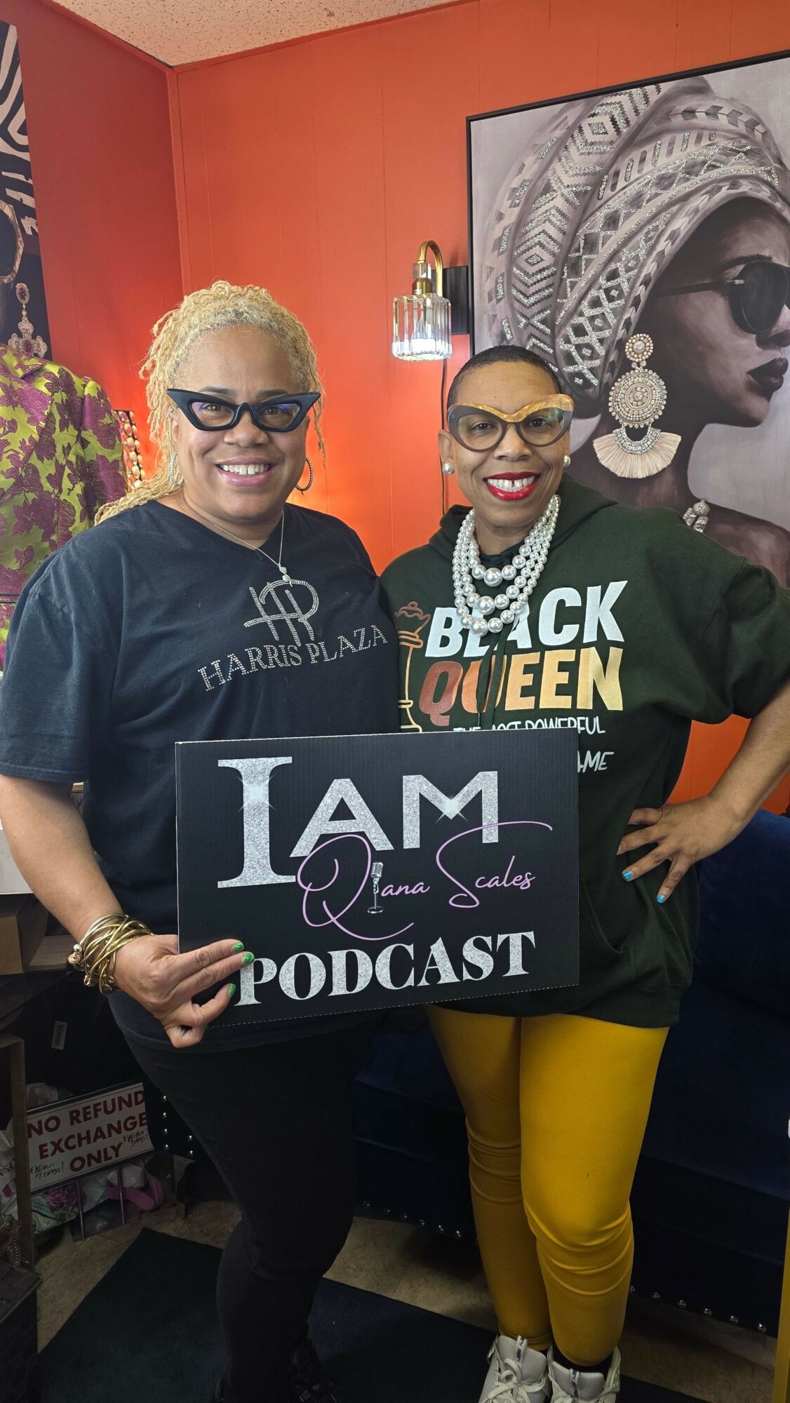 Two women holding a sign that reads 'I AM PODCAST' in a room with orange walls and artwork.
