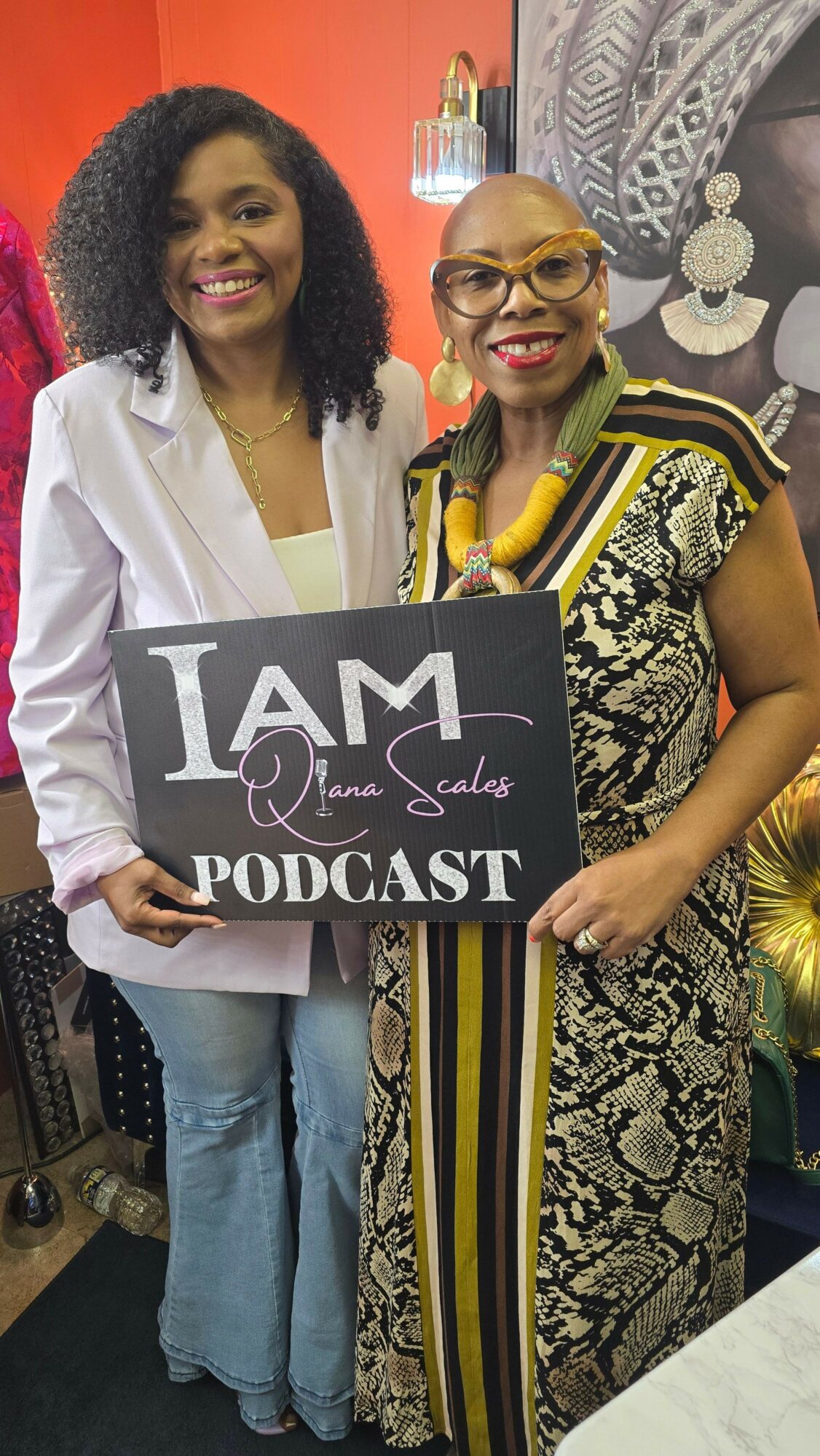 Two women standing indoors, one holding a sign that reads 'I AM Podcast,' smiling at the camera.