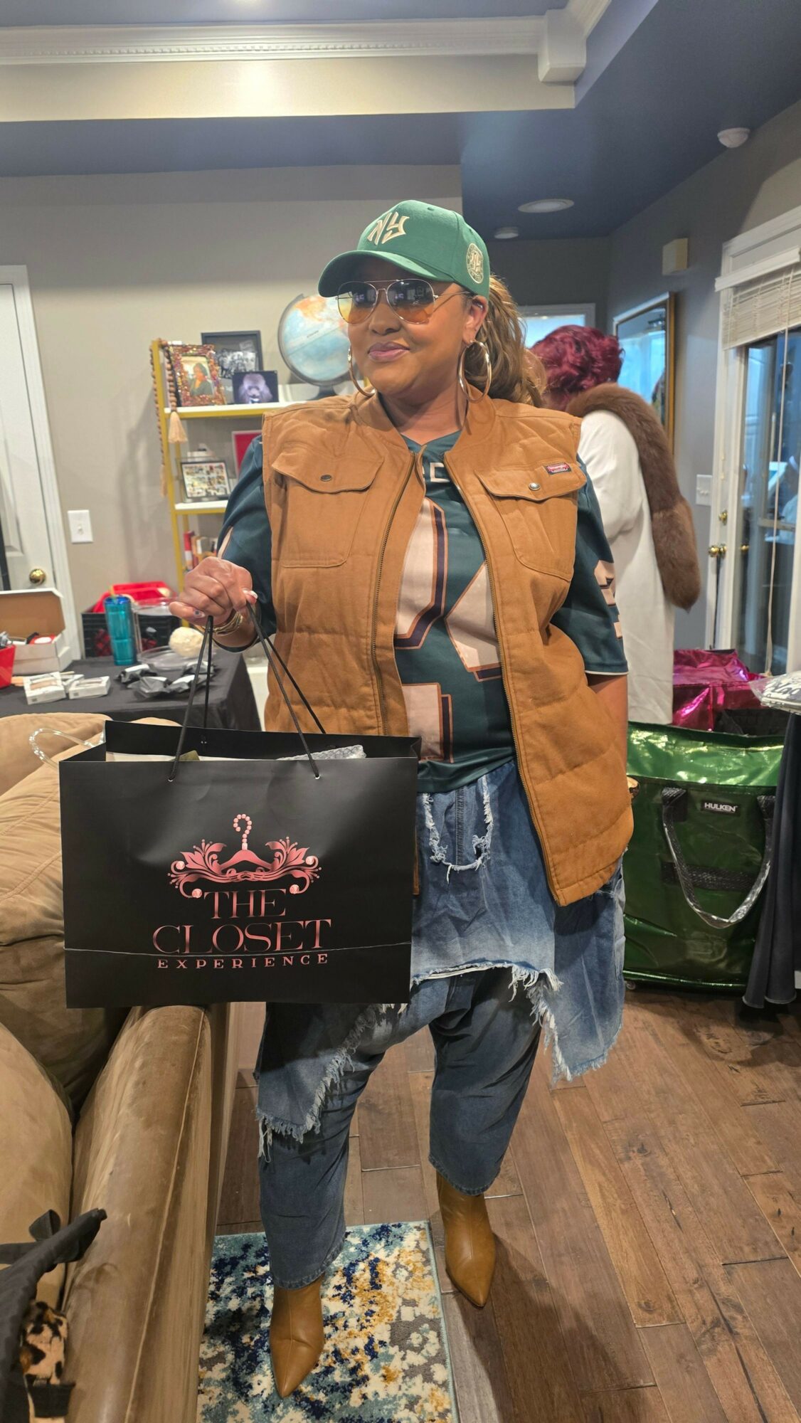 Person with a brown jacket, jeans, and a green cap holding a shopping bag in a store, with shelves and other shoppers visible.