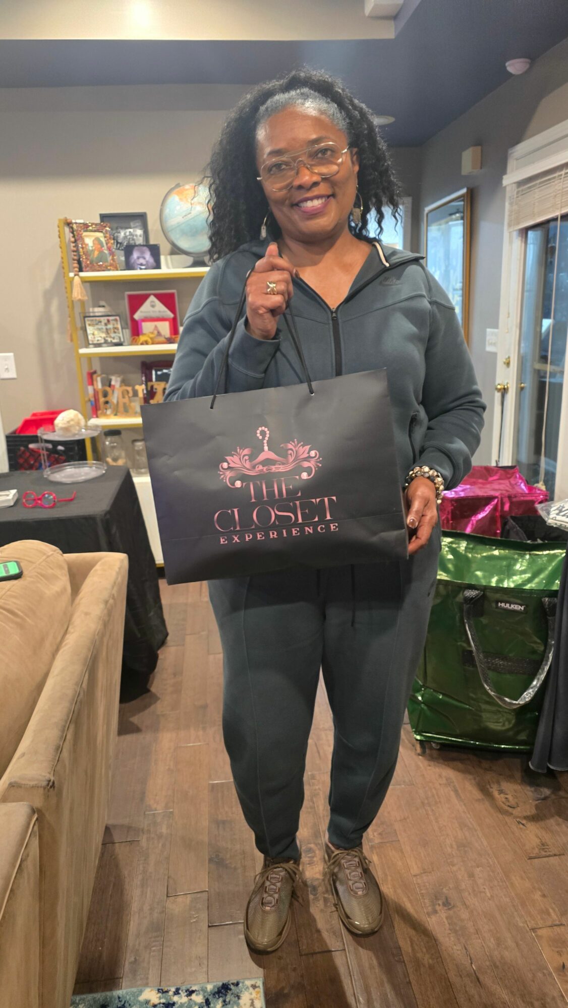 Woman standing indoors holding a black bag with pink text, smiling, with a green suitcase and various items in the background.