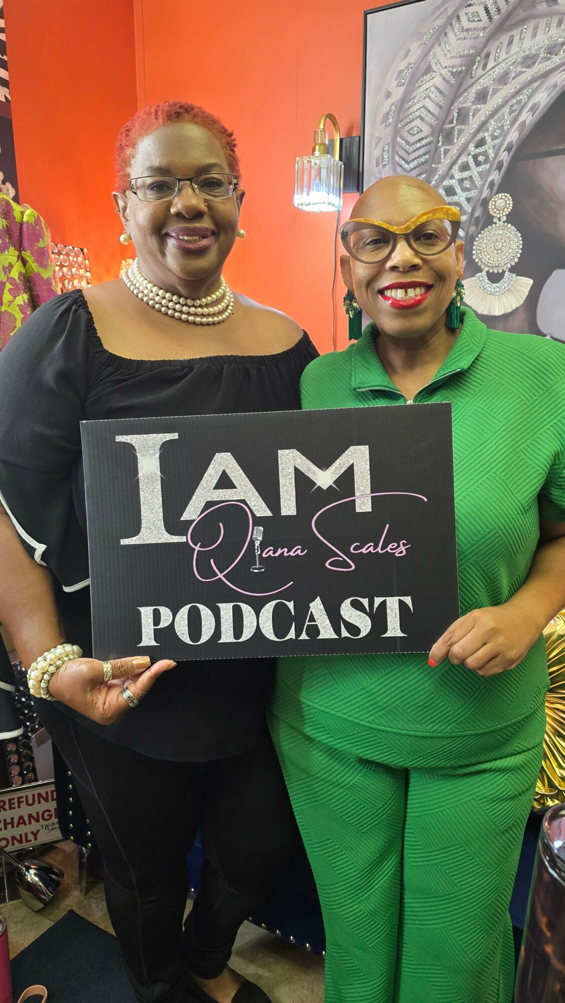 Two women smiling, one holding a sign that says 'I AM Podcast,' in a colorful indoor setting.