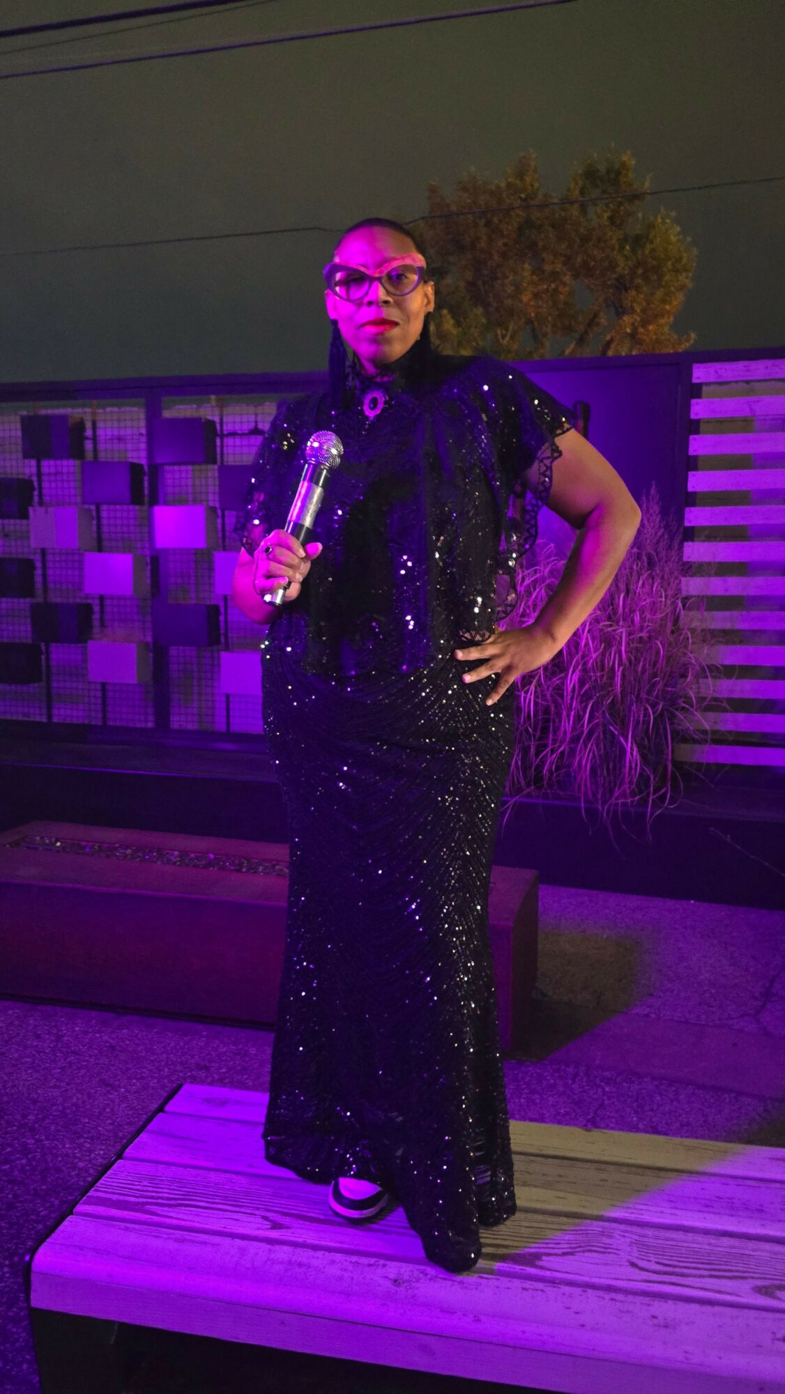 Person in a black, sparkly dress holding a microphone, standing on a wooden floor with purple lighting.