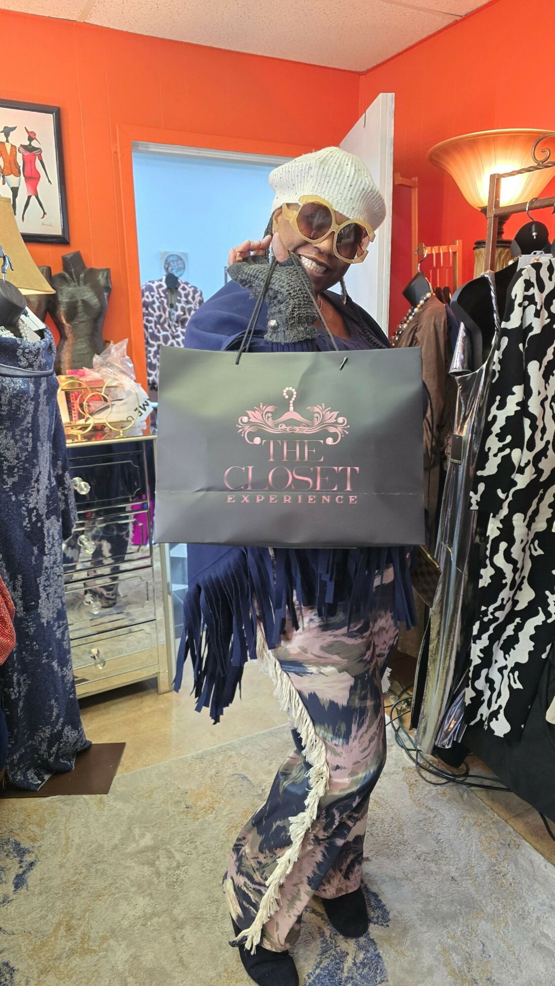 Person dressed in camouflage clothing and sunglasses holding a sign with a pink flamingo, standing indoors with orange walls.