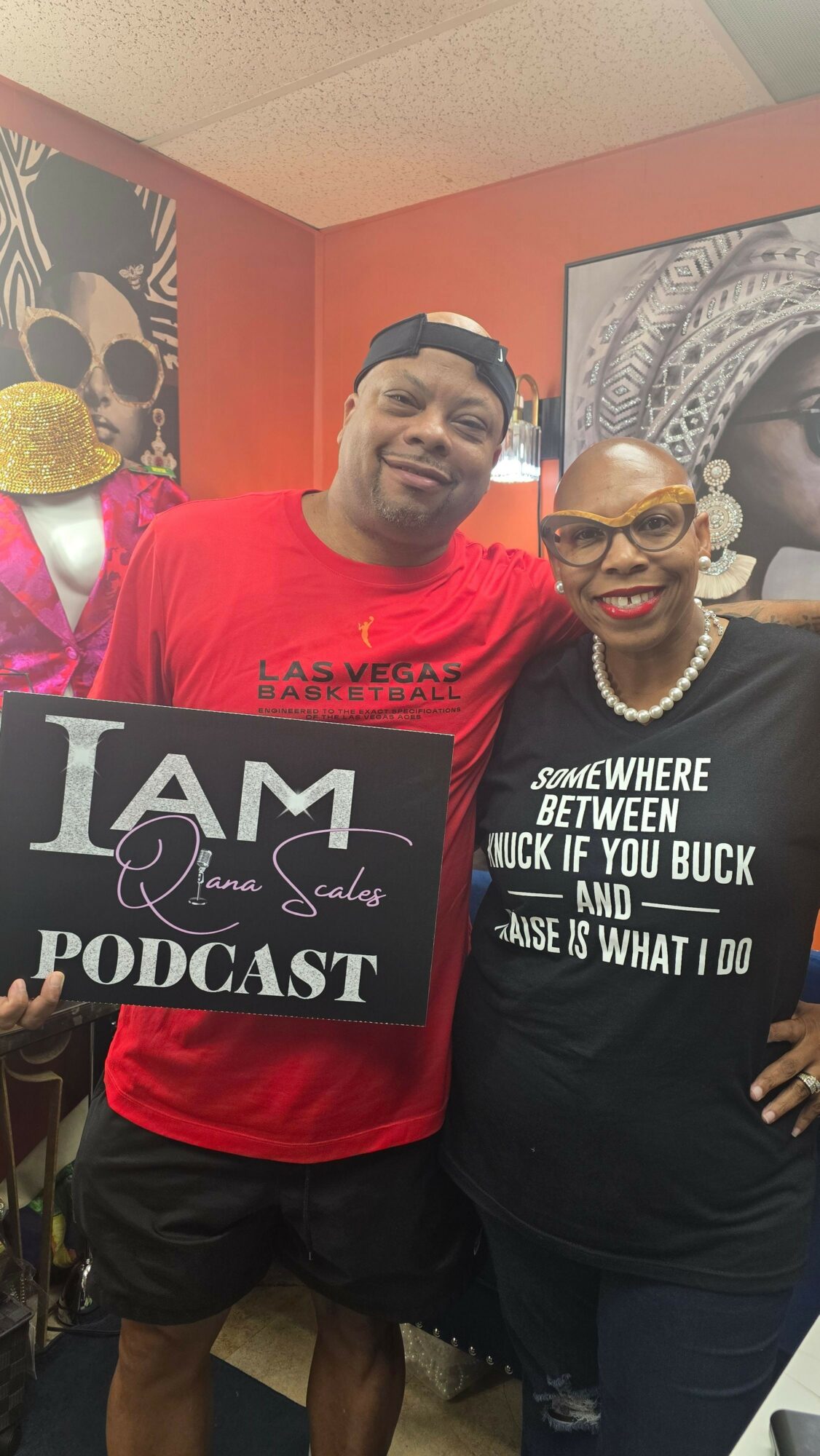 Two men standing indoors, one holding a podcast sign, both smiling, with artwork on the wall behind them.