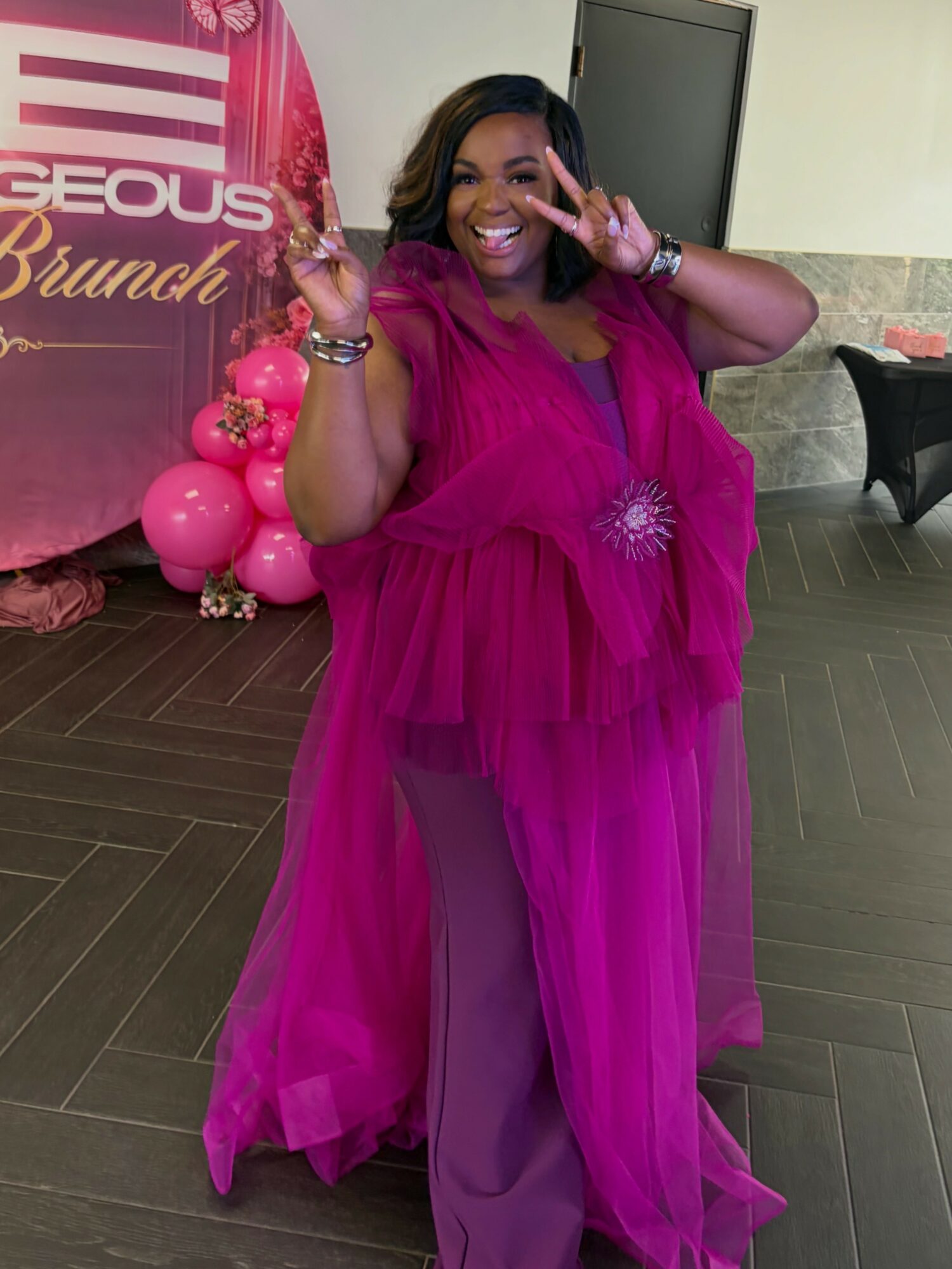 Smiling woman in pink dress making peace signs, standing indoors near pink balloons and a banner.