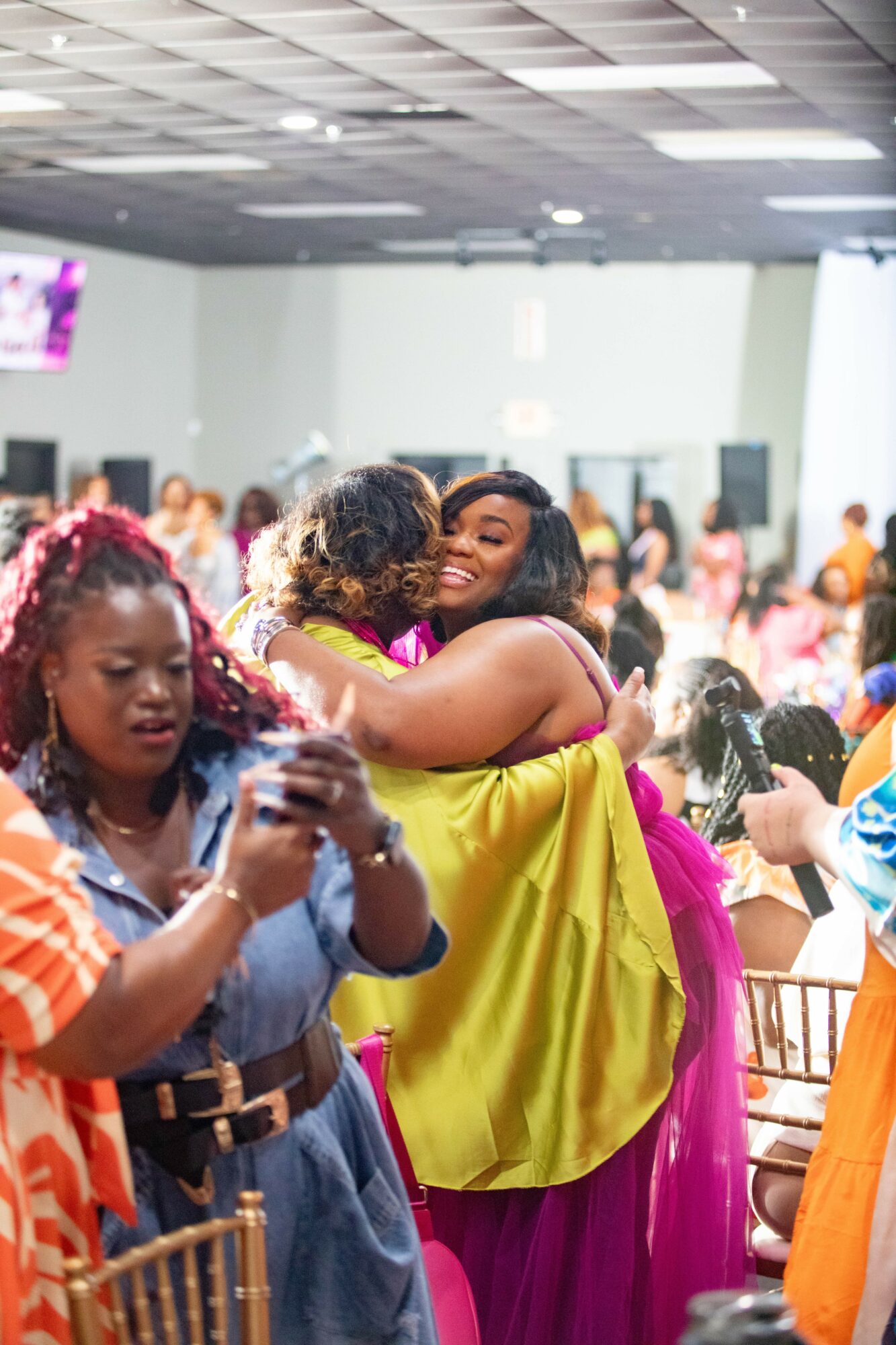 Two women hug in a crowded indoor event, with other people in the background, some taking photos.