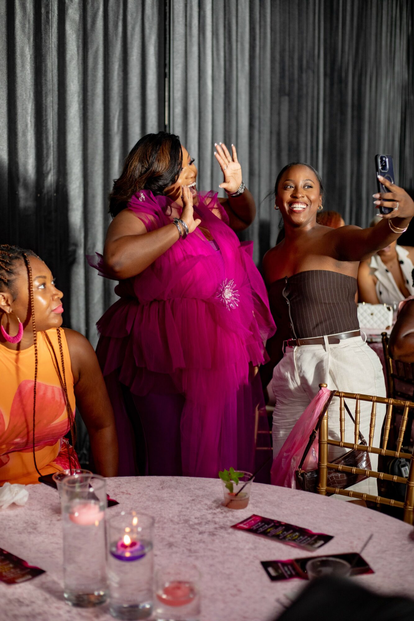 Three women at a celebration, one in a pink dress, two smiling and taking photos, with a black curtain background.