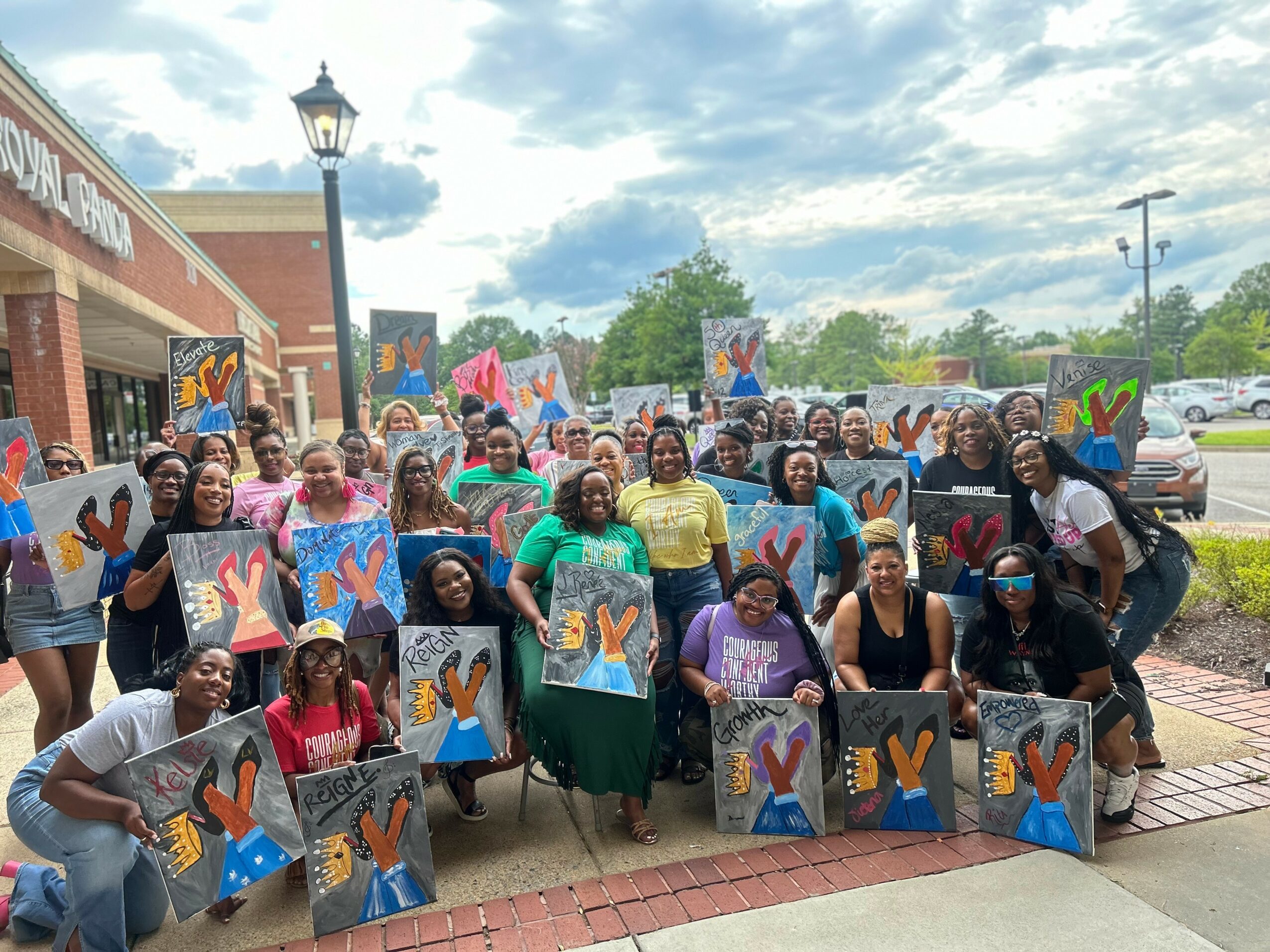 Group of people outdoors holding colorful paintings with handprints, smiling, in front of a building and parked cars.