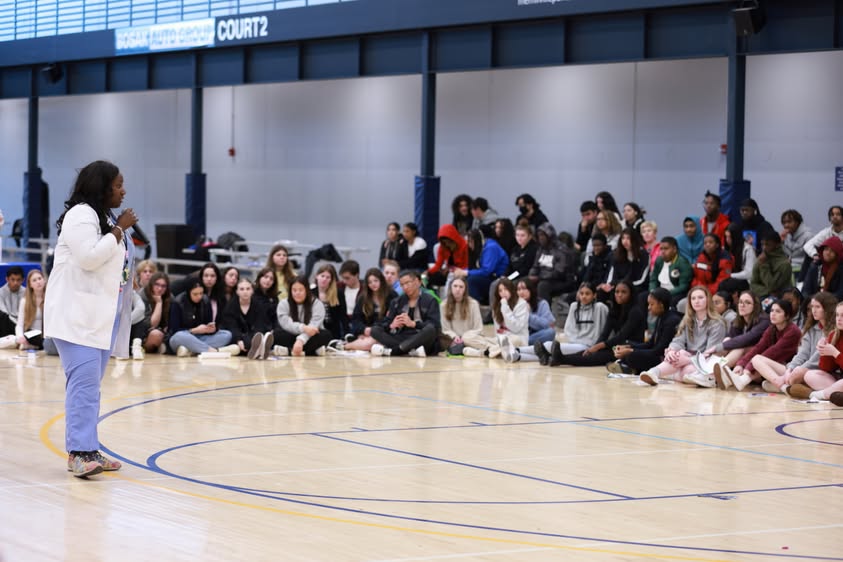 A woman speaking to a large group of people seated on the floor in a gymnasium.