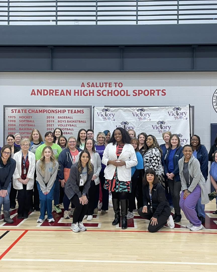 Group of people posing in a gymnasium with a banner honoring Andrean High School sports, and a list of state championship teams.