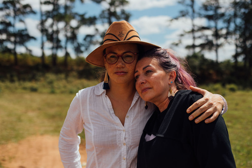 Two women outdoors, one wearing a hat and glasses, the other with dark hair, hugging each other.