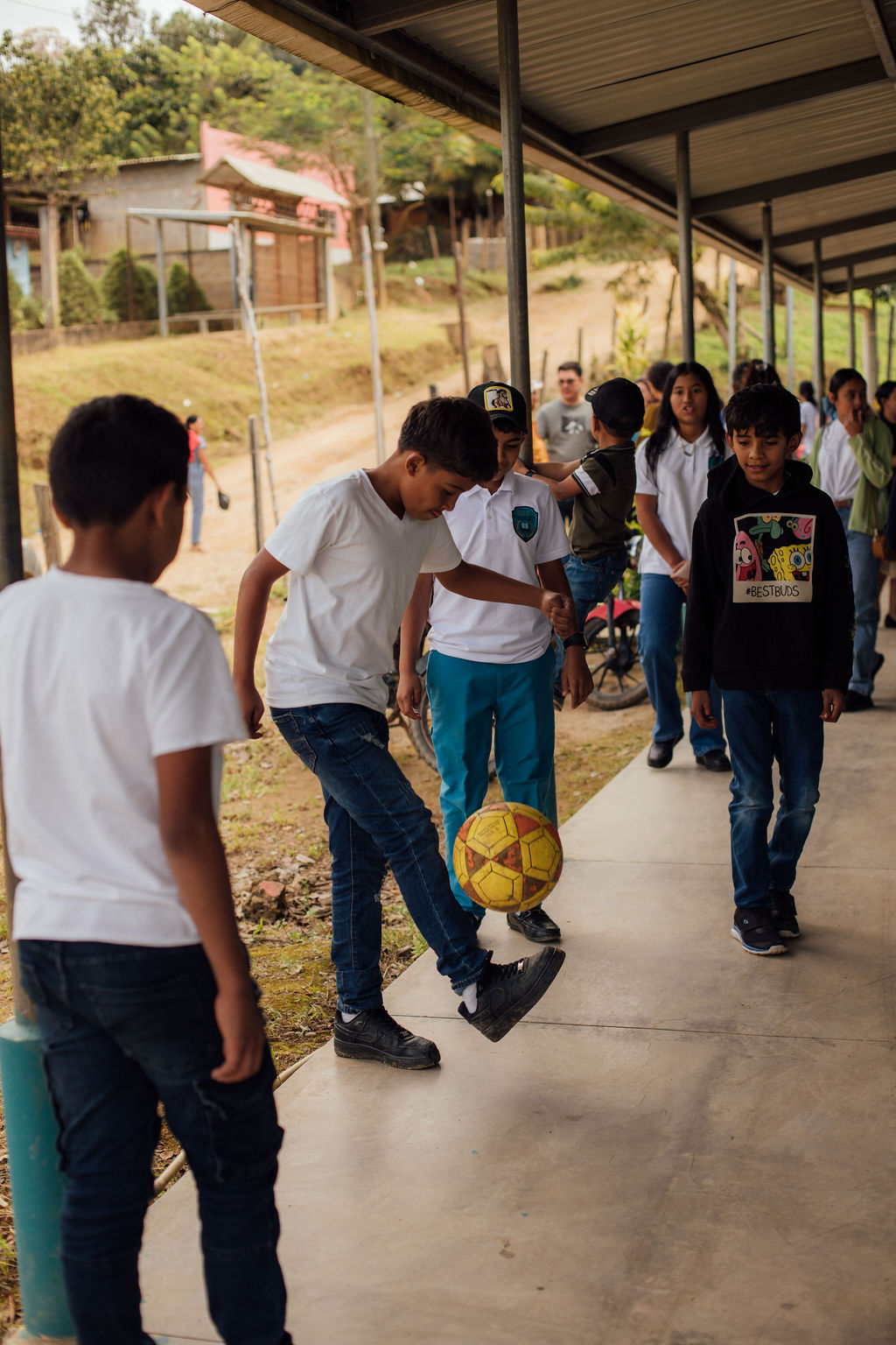 Group of children walking and playing with a yellow soccer ball under a covered walkway outdoors.