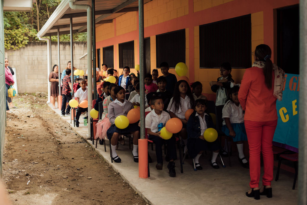 Children sitting outdoors with balloons, some standing and some sitting, near a colorful building, with a woman in red nearby.