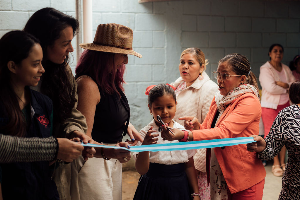 Group of women and children holding a ribbon at a celebration indoors, with a brick wall background.