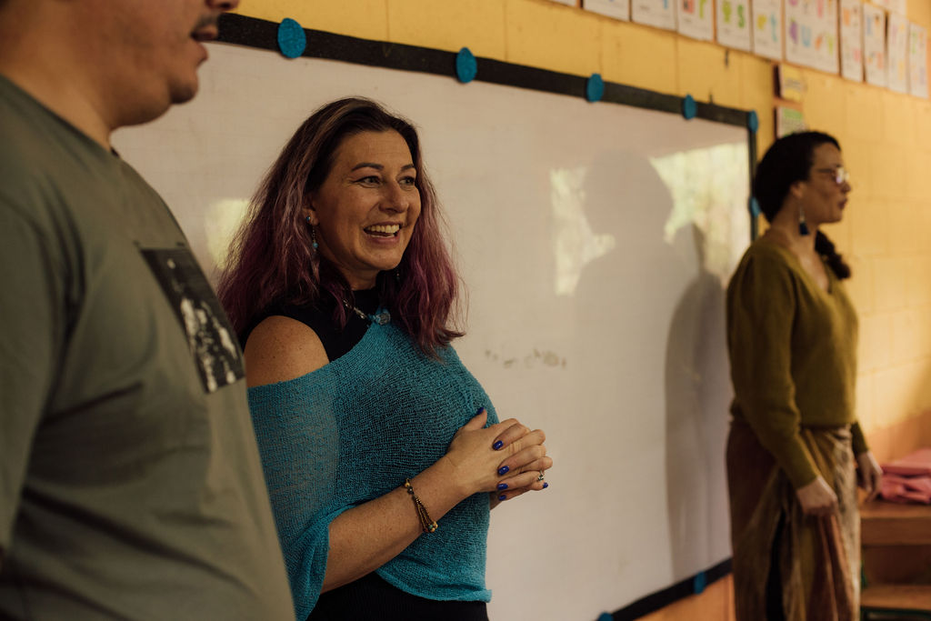 Three people in a classroom, woman smiling and clapping, whiteboard behind them, other two women standing nearby.