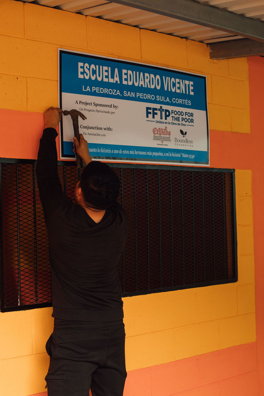 Person hanging a sign on a yellow wall with a blue school sign above a blackboard, indoors.