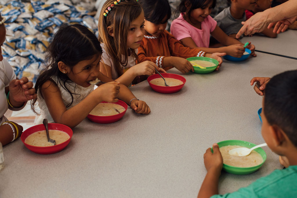 Children sitting at a table with colorful bowls, reaching for food or objects, engaged in an activity.