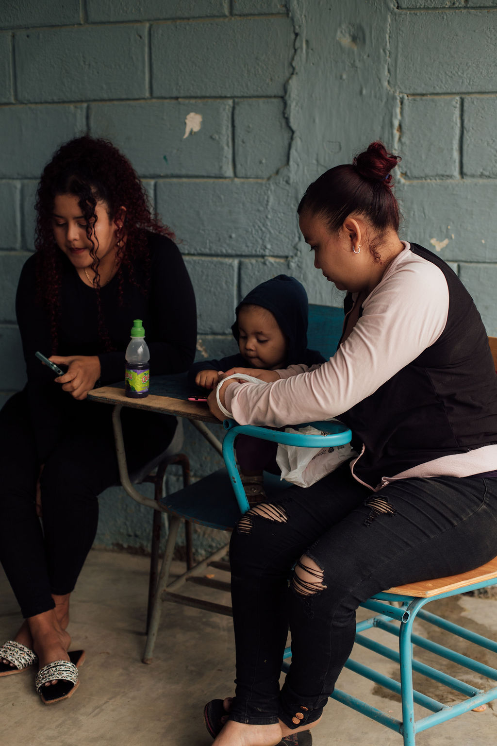 Two women and a child sitting on chairs against a brick wall, with a small table and a bottle nearby.