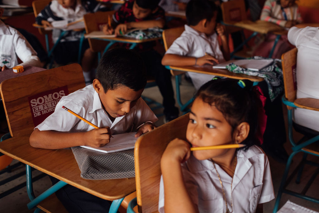 Children sitting at desks in a classroom, writing and engaging in activities, wearing white uniforms.