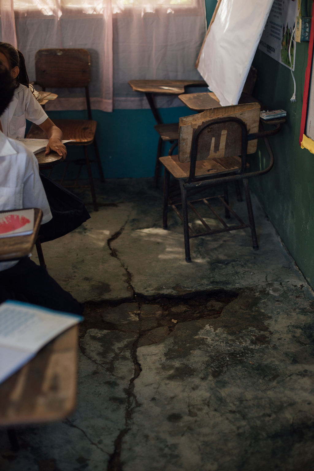 Classroom with cracked floor, desks, and chairs, with a person sitting at a desk, and a window with curtains in the background.