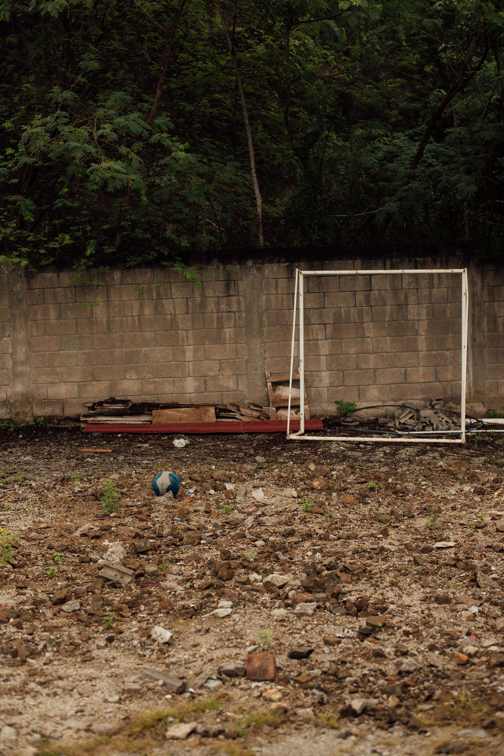 Empty soccer goal on a dirt field with a brick wall and trees in the background.